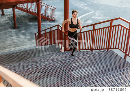 Young athletic slim beautiful woman running up stairs doing cardio interval training in black fashionable sportwear in the street on summer. Outdoor, sport and healthy concept Young athletic slim beautiful woman running up stairs doing cardio interval training in black fashionable sportwear in the street on summer. Outdoor, sport and healthy concept 88711865