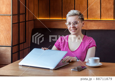 Portrait of beautiful satisfied happy young bussineswoman with blonde short hair in pink t-shirt is sitting in cafe, finishing work and looking at camera with toothy smile. Indoor, healthy lifestyle Portrait of beautiful satisfied happy young bussineswoman with blonde short hair in pink t-shirt is sitting in cafe, finishing work and looking at camera with toothy smile. Indoor, healthy lifestyle 88711879