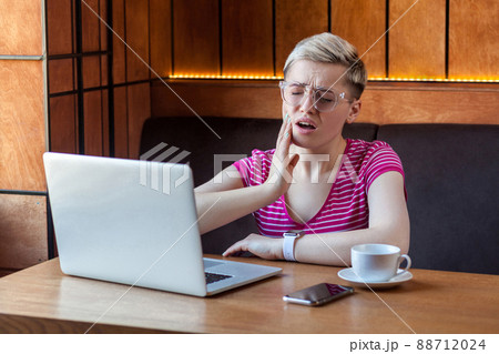 Portrait of unwell young adult woman with short blonde hair in pink t-shirt and eyeglasses sitting in cafe and touching cheek with hand have a high pain, toothache and dentistry. Indoor, health care 88712024