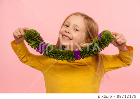 Portrait of happy preschool child wearing green wreath around neck and smiling broadly to camera, having fun, showing hawaii necklace, masquerade accessory. studio shot isolated on pink background Portrait of happy preschool child wearing green wreath around neck and smiling broadly to camera, having fun, showing hawaii necklace, masquerade accessory. studio shot isolated on pink background 88712179