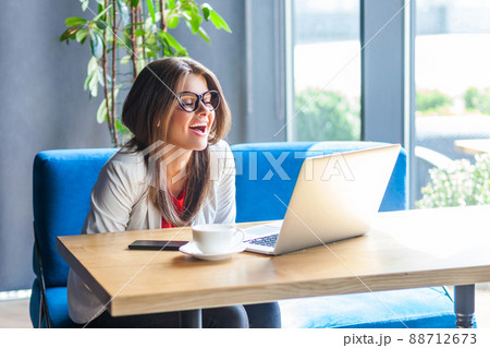 Portrait of joyful beautiful stylish brunette young woman in glasses sitting, looking at her laptop screen on video call and laughing or amazed. indoor studio shot, cafe, office background. 88712673
