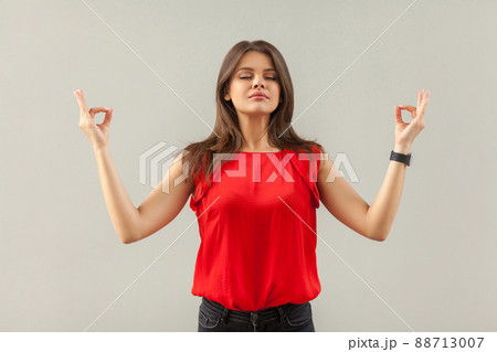 Yoga and relaxation. Portrait of calm beautiful brunette young woman in red shirt standing in yoga pose with closed eyes and meditating. indoor, studio shot, isolated on gray background. 88713007