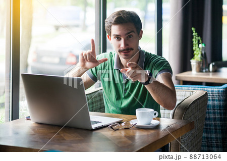 You are loser. Young serious businessman in green t-shirt sitting, pointing and showing loser gesture and looking at camera. business and freelancing concept. indoor shot near big window at daytime. 88713064