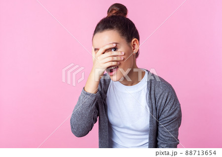 Portrait of adorable curious brunette teen girl with bun hairstyle in casual clothes peeking through fingers, looking embarrassed, pleasantly surprised. indoor studio shot isolated on pink background Portrait of adorable curious brunette teen girl with bun hairstyle in casual clothes peeking through fingers, looking embarrassed, pleasantly surprised. indoor studio shot isolated on pink background 88713654