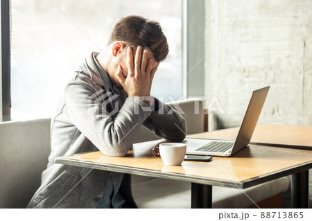 Portrait of sad tired alone depressed tired bearded young freelancer in grey blazer are sitting alone in cafe and searching for a work on laptop are in bad mood without good result. 88713865