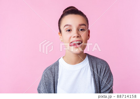 Portrait of carefree childish brunette girl with bun hairstyle in casual clothes showing tongue, disobedient teenager making face, having fun at camera. indoor studio shot isolated on pink background Portrait of carefree childish brunette girl with bun hairstyle in casual clothes showing tongue, disobedient teenager making face, having fun at camera. indoor studio shot isolated on pink background 88714158