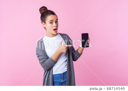 Wow, look at mobile app. Portrait of surprised teenage brunette girl with bun hairstyle in casual clothes pointing at cellphone, amazed shocked about gadget. studio shot isolated on pink background 88714540