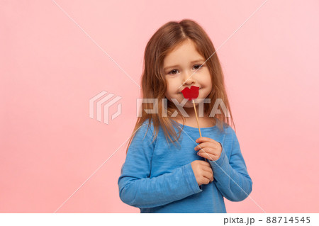 Portrait of lovely cute brunette little girl covering mouth with red paper lips or heart shape, kindly looking at camera, fashion and beauty concept. indoor studio shot isolated on pink background Portrait of lovely cute brunette little girl covering mouth with red paper lips or heart shape, kindly looking at camera, fashion and beauty concept. indoor studio shot isolated on pink background 88714545