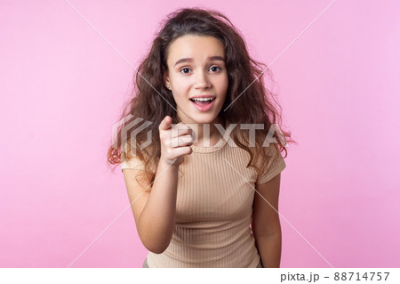 Hey you. Portrait of surprised teen girl with curly brunette hair wearing casual style beige clothes pointing at camera, standing with open mouth, wondered astonished expression. studio shot, indoor 88714757
