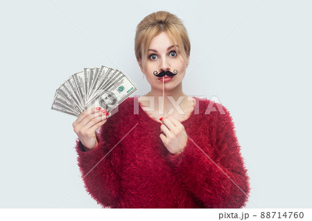 Beautiful young woman in red blouse standing, demonstrate fan of money, holding manlike mustache sticker, looking at camera with big eyes. Indoor, studio shot, isolated on gray background 88714760