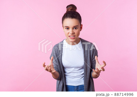 Portrait of irritated brunette girl with bun hairstyle in casual clothes raising hands, looking at camera with mad face, clenched teeth, rebellious teenager. studio shot isolated on pink background 88714996
