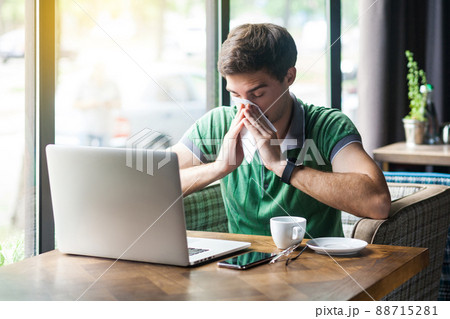 Young cold sick businessman in green t-shirt sitting at work, sneezing and cleaning his nose with tissue napkin. business and helthcare concept. indoor shot near big window at daytime. 88715281