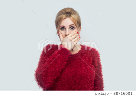 Portrait of shocked silence beautiful young blond woman in red blouse standing, covering her mouth and looking at camera with big eyes. indoor studio shot isolated on light gray background. 88716001