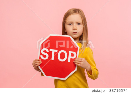 Portrait of cute little girl holding Stop symbol, red traffic sign and looking with serious responsible expression, warning about road safety rules. indoor studio shot isolated on pink background Portrait of cute little girl holding Stop symbol, red traffic sign and looking with serious responsible expression, warning about road safety rules. indoor studio shot isolated on pink background 88716574
