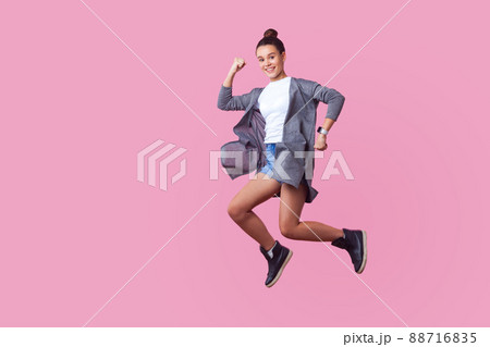 Ecstatic brunette teenage girl with bun hairstyle in casual clothes jumping in the air with enthusiastic happy face, raised hands and running in hurry. indoor studio shot isolated on pink background Ecstatic brunette teenage girl with bun hairstyle in casual clothes jumping in the air with enthusiastic happy face, raised hands and running in hurry. indoor studio shot isolated on pink background 88716835