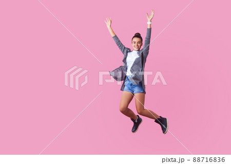Excited energetic brunette teenage girl with bun hairstyle in casual clothes jumping in the air with enthusiastic happy face and raised hands, optimistic life. studio shot isolated on pink background 88716836
