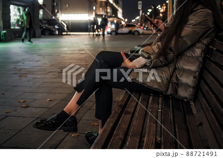 Beautiful elegant women's legs in black glossy loafers. A woman sits on a bench on a night street in the city in autumn 88721491