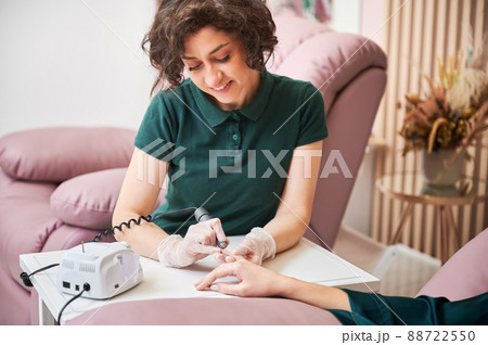 Female manicure master sitting at the table and smiling while doing hardware manicure on woman hand. Manicurist removing cuticle on client hand with electric nail drill. 88722550