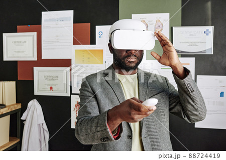 African medical specialist in virtual reality goggles having virtual conference while standing at office African medical specialist in virtual reality goggles having virtual conference while standing at office 88724419