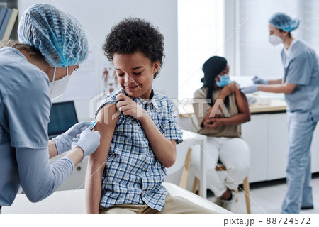 African brave boy getting vaccination in shoulder from nurse during procedure at hospital with other people in background 88724572