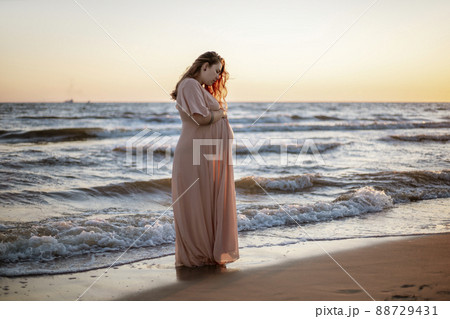 Young pregnant woman with a beautiful sea view on the background. Happy and calm pregnant woman with long hair and pink dress standig on the beach. Romantic view, ocean, sunset, maternity. Young pregnant woman with a beautiful sea view on the background. Happy and calm pregnant woman with long hair and pink dress standig on the beach. Romantic view, ocean, sunset, maternity. 88729431