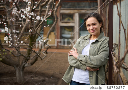 Confident middle aged Caucasian woman gardener enjoying an early spring day standing in her garden plot against the blossoming apricot fruit trees background Confident middle aged Caucasian woman gardener enjoying an early spring day standing in her garden plot against the blossoming apricot fruit trees background 88730877