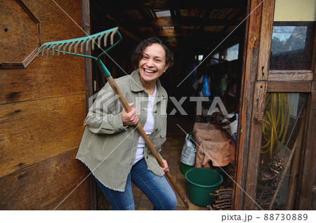 Happy pleasant mature female gardener holding a rake and smiling with a cheerful toothy smile while standing in her wooden rural workshop with gardening tools enjoying home gardening on a spring day 88730889