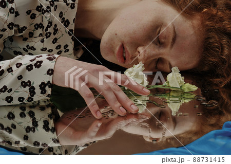 Young woman lying with her eyes closed and touching the flower lying on wet mirror 88731415