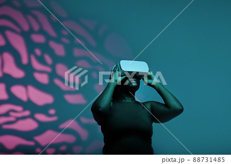 African plump woman in headset playing virtual reality standing against the pink and blue background 88731485