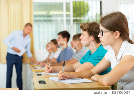 Teen girl in glasses listening to lecturer and writing in notebook in classroom 88736034