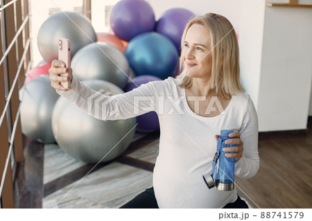 Caucasian pregnant woman in third trimester make a selfie and holding a bottle with water. Woman in bright fitness studio with big windows. Blonde woman wearing whit shirt and black leggins. 88741579