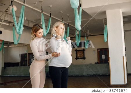 Pregnant woman doing yoga with personal trainer. Yoga trainer assisting pregnant woman while doing exercises with a fitball and dumbbells. Blonde pregnant woman wearing white shirt. 88741674