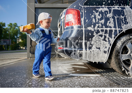 Kid boy studying work at car wash. Toddler child, holding yellow foam sponge in his right hand, examining soap car and preparing to start cleaning the back part of transport. Kid boy studying work at car wash. Toddler child, holding yellow foam sponge in his right hand, examining soap car and preparing to start cleaning the back part of transport. 88747825