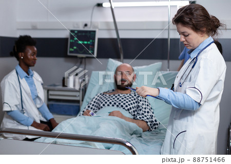 Middle aged man recovering lying in hospital bed attended by team of doctors after surgical intervention. Doctor with stethoscope reading medical chart while nurse is monitoring patient vitals. 88751466