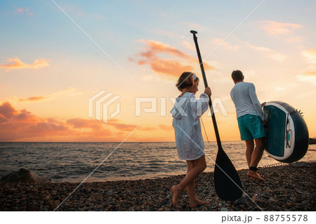 Summer vacations. Father and daughter are walking along the beach with a sup board. Sea and the sunset in the background. Back view. Sea surfing 88755578