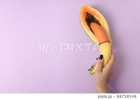 Female hand holding banana with condom in papaya on violet background Female hand holding banana with condom in papaya on violet background 88758548