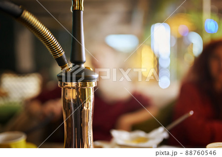Hookah on table in lounge bar. Close-up of black pipe and golden shisha flask. Smoking person releases thick fume in blurry background. Leisure in oriental cafe. 88760546