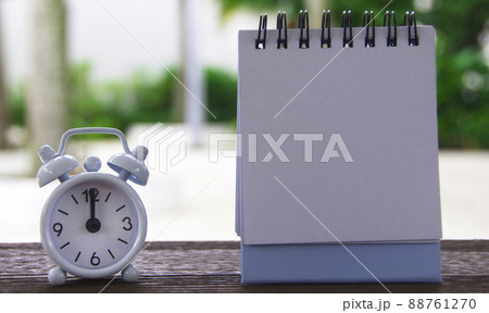 Alarm clock pointing at 12 o'clock with blank notepad. Blurred park background. Copy space 88761270