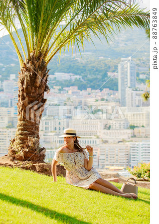 a bright beautiful girl in a light dress and hat sits on the grass under a palm tree in Monaco in sunny weather in summer, streets of old town of Monaco 88763849
