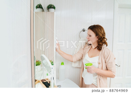 an attractive housewife washes the bathroom mirror with a special glass cleaner. an attractive housewife washes the bathroom mirror with a special glass cleaner. 88764969