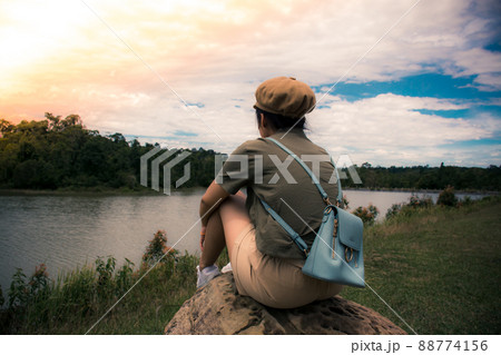 Young asian lonely girl while sitting at stone next to the river among mountains. She was wearing a green shirt & shoulder bag, looking at the sunset and thinking about her problems and felt very sad. Young asian lonely girl while sitting at stone next to the river among mountains. She was wearing a green shirt & shoulder bag, looking at the sunset and thinking about her problems and felt very sad. 88774156