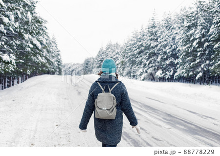 a girl in a gray coat with a gray backpack walking the side of a winter road, next to a winter forest of pine trees 88778229