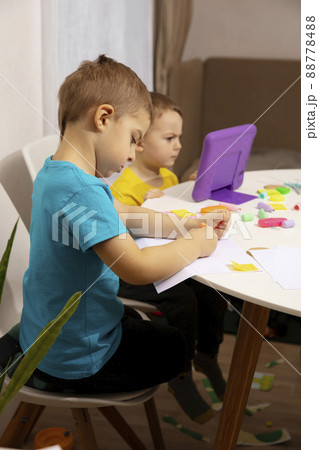 Happy kids with yellow and blue shirts doing arts and crafts together at their desk and watching cartoon on digital tablet. Children have fun. Boys play at home. 88778488