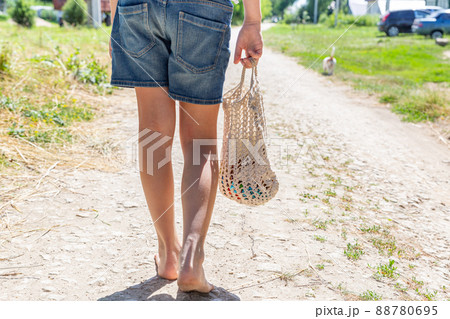 The girl goes to the store barefoot along the village road, bare feet, rear view. Eco-friendly reusable mesh bag, handmade, macrame. Eco-friendly concept without waste. 88780695