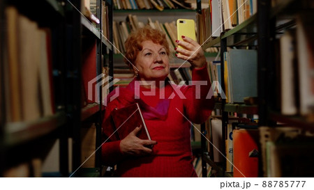 An elderly woman takes a photo with books in the library. Selfie for social networks 88785777