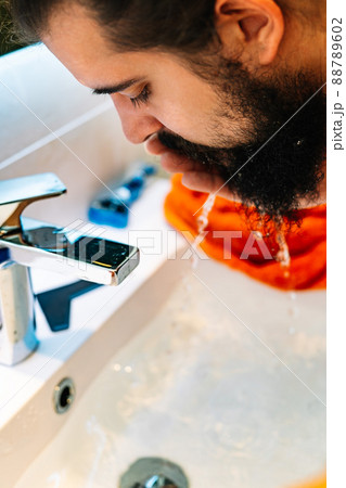 close-up of a young man with bushy beard, washing his face in the bathroom sink 88789602
