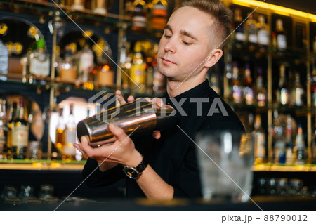 Close-up of bartender in black clothes mixing ingredients of alcoholic cocktail by shaking shaker standing behind bar counter, on blurred background of shelves with alcoholic drinks, selective focus. 88790012