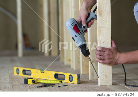 Close-up of worker's hands with screwdriver on background of professional tools and wooden frame for future wall in unfinished attic room under reconstruction. Renovation and improvement concept. 88794504