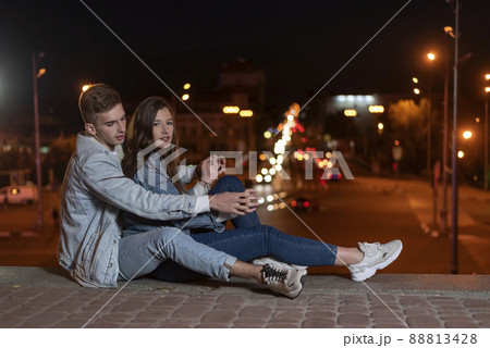 Young caucasian couple is sitting on the pavement hugging each other against the backdrop of the lights of the night city. Date and relationship. Side view Young caucasian couple is sitting on the pavement hugging each other against the backdrop of the lights of the night city. Date and relationship. Side view 88813428