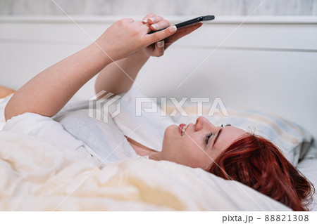 young woman smiling, taking a selfie with cell phone, lying on her bedroom bed. 88821308